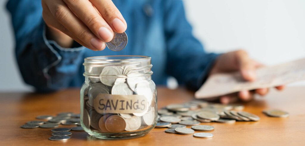 Person dropping coins into small glass jar marked "Savings".