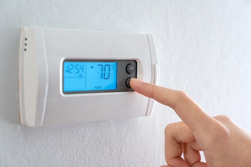 A woman is pressing the down button of a wall attached house thermostat with digital display showing temperature 70 degree Fahrenheit for heating, cooling, electricity and gas saving