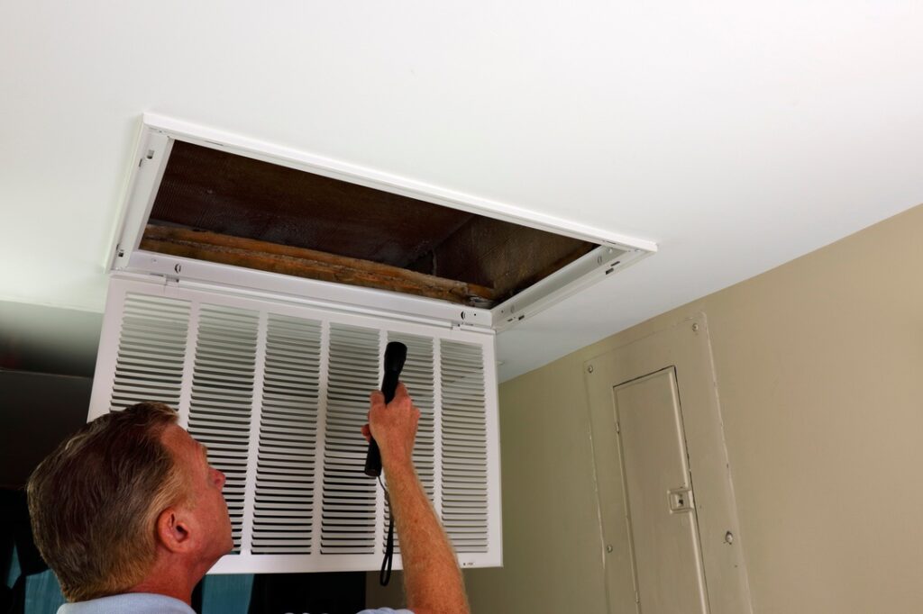 Flashlight shined into an air intake vent of a home HVAC system by an adult white male in an entryway. Adult male looking into an air intake vent with open grid after removing home HVAC air filter