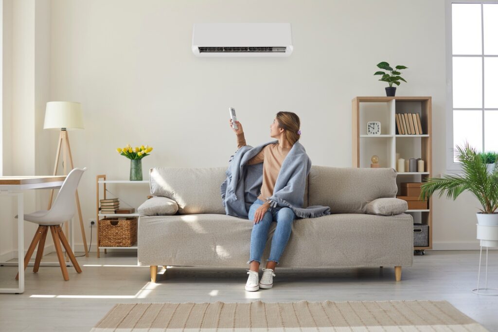 Woman who's sitting on sofa under warm plaid in living room switches off her air conditioner on wall. Young girl adjusting modern AC system, regulating temperature and enjoying cool fresh air at home